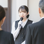 女性司会者がマイクで話す様子が写された、葬儀の進行中の場面