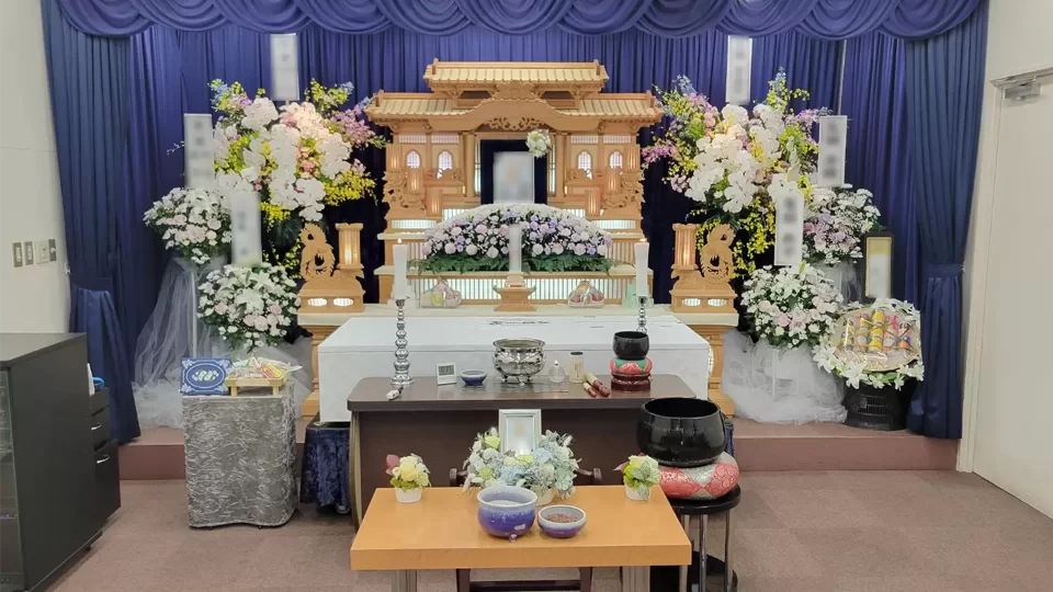 背景に青い幕を配し、中央に白木祭壇と多くの花が並ぶ整然とした会場